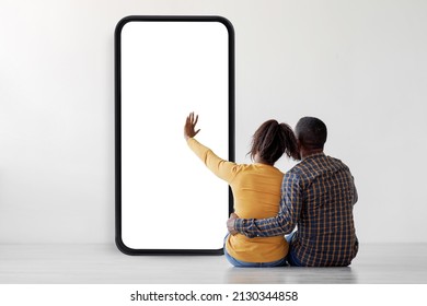 Back view of African American couple looking at huge smartphone with blank screen, sitting on floor against white studio wall, using new mobile app or website, checking online ad, mockup