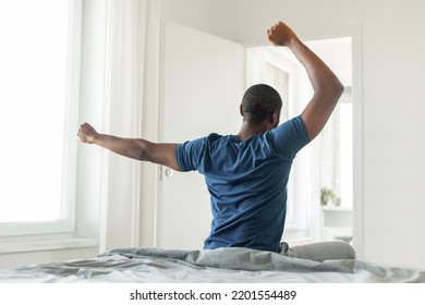 Back View Of African American Guy Stretching Arms Sitting On Comfortable Bed After Good Sleep In Moderrn Bedroom At Home. Healthy Morning Routine Concept