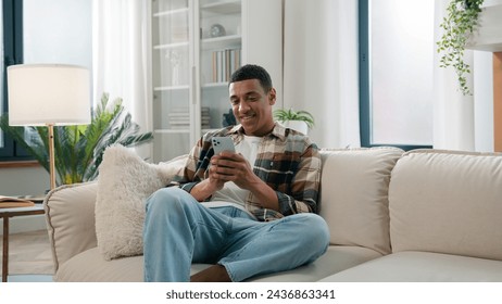 Back view African American guy enter living room walk at home approach go to sofa sit on couch browsing mobile phone man scrolling smartphone social media indoors using cellphone app chatting relaxing
