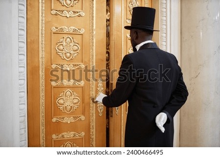 Back view of African American lord wearing top hat opening carved wooden doors and entering mansion, copy space