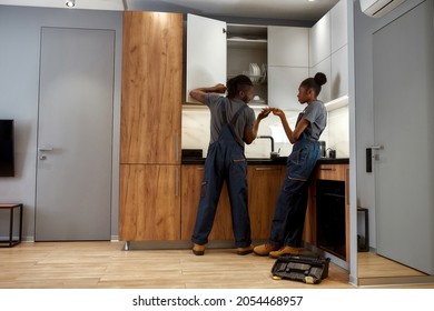 Back View African American Man And Woman, Repair Service Workers, Fixing Cupboard In Modern Kitchen. Skilled Handyman Taking Tool From Demotivated Female Apprentice.