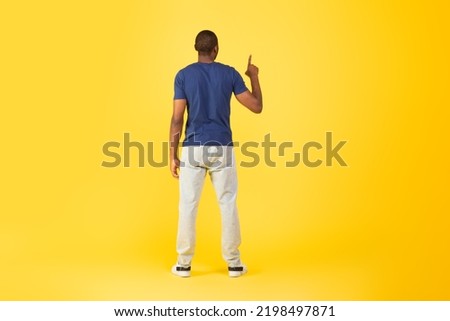 Back View Of African American Man Pointing Finger Up Pushing Invisible Button Or Touchscreen Standing Over Yellow Studio Background. Look Upward. Full Length Shot