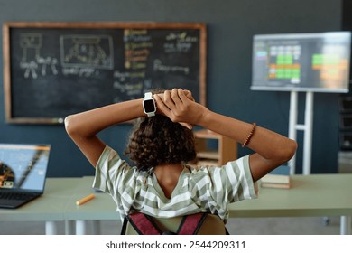 Back view of African American teenage girl stretching while sitting at desk in school classroom with smartwatch in focus wearable tech copy space - Powered by Shutterstock