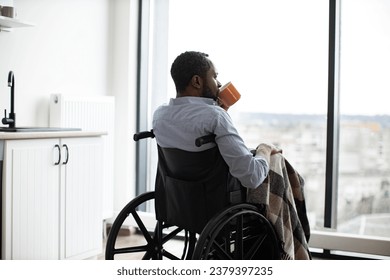 Back view of african american wheelchair user with cup of hot drink looking at panoramic window resting in open-plan kitchen of modern apartment. - Powered by Shutterstock