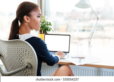 Back View Of African Business Woman In Dress Sitting By The Table Near The Window In Office With Laptop And Looking Aside