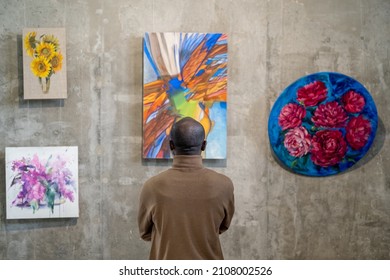 Back view of African male guest of art gallery standing in front of wall with expositions and looking at one of them - Shutterstock ID 2108002526
