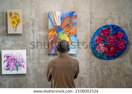 Back view of African male guest of art gallery standing in front of wall with expositions and looking at one of them