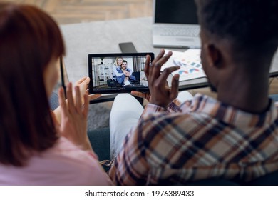 Back View Of African Man And Caucasian Woman Waving Hand During Video Conversation With Relatives. Young Couple Using Digital Tablet For Online Communication At Home.