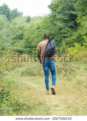 A back view of an African tall man in stylish clothes with a backpack walking on the grassy pathway