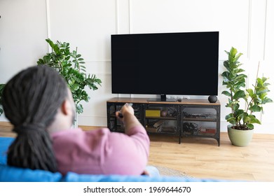 Back View African-American Guy With Dreadlocks Holds Remote Controller And Switching Tv Channels, Watching TV Sitting On The Couch At Home, The Lcd TV With Empty Black Screen, Copy Space, Mockup