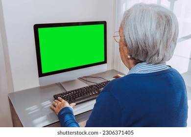 Back view of aged woman, senior old mature grandmother looking focused using pc, sitting at desk, surfing internet. Mock-up green screen. - Powered by Shutterstock