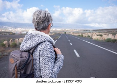 Back View Of Alone Senior Woman Walking Outdoor Wearing Backpack Looking At Mountain Landscape. Elderly Mature Lady In Empty Road, Mountain And Blue Sky On Background