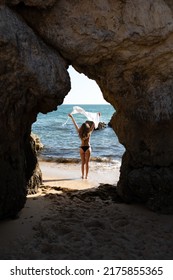 Back View Of Anonymous Distant Female Traveler In Bikini Standing On Sandy Beach With Rocky Formation While Admiring Rippling Sea