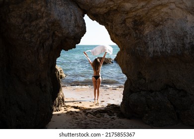 Back View Of Anonymous Distant Female Traveler In Bikini Standing On Sandy Beach With Rocky Formation While Admiring Rippling Sea