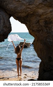 Back View Of Anonymous Distant Female Traveler In Bikini Standing On Sandy Beach With Rocky Formation While Admiring Rippling Sea