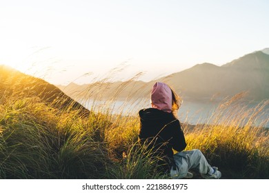 Back View Of Anonymous Female Traveler In Warm Hoodie Sitting On Grassy Hill Slope And Admiring Mountainous Lake Against Cloudless Sunset Sky During Trip In Mount Batur In Bali