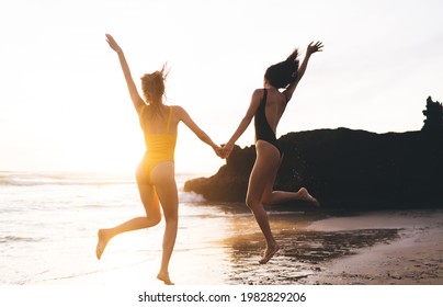 Back view of anonymous girlfriends holding hands while jumping on sandy shore near calm rippling water of ocean and cliff - Powered by Shutterstock