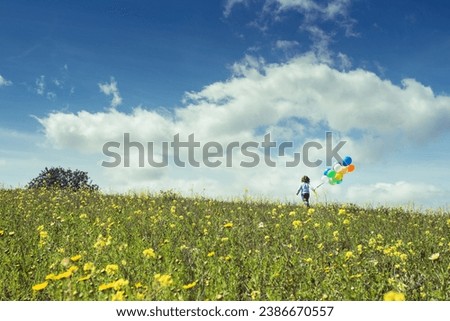 Back view of anonymous kid holding colorful balloons while walking on blooming meadow under cloudy sky on sunny day