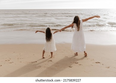 Back View Of Anonymous Mom And Kid In White Dresses Spreading Arms And Enjoying Freedom While Spending Time On Sandy Beach Near Waving Sea