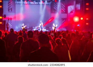 Back view of anonymous spectators looking away while standing near stage with blurred musicians playing instruments and performing in live concert in illuminated pink neon spotlights