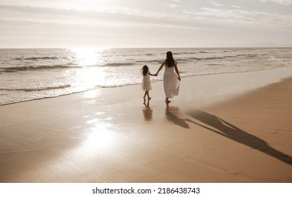 Back View Of Anonymous Woman And Preteen Girl With Long Dark Hair In Similar White Dresses, Holding Hands And Strolling On Wide Sandy Beach Near Wavy Ocean Against Sunset Sky