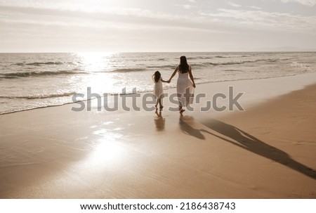 Back view of anonymous woman and preteen girl with long dark hair in similar white dresses, holding hands and strolling on wide sandy beach near wavy ocean against sunset sky