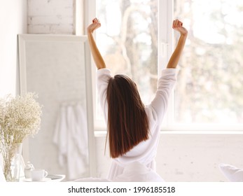 Back View Of Anonymous Woman In Sleepwear Raising Arms And Stretching Body While Sitting On Bed In Morning In Light Bedroom