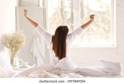 Back View Of Anonymous Woman In Sleepwear Raising Arms And Stretching Body While Sitting On Bed In Morning In Light Bedroom