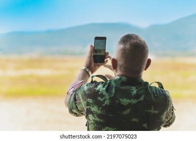 Back View Of Asian Army Soldier Using Cell Phone To Take Landscape Photo In Field. Taking Photo On Mobile Phone Of Inspect The Battlefield. Combatant Takes Picture And Check Maneuver With Smartphone.