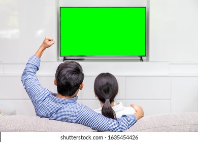 Back View Of Asian Couple Sitting On Sofa Cheering And Enjoy Watching Sport On TV At Home With Green Screen