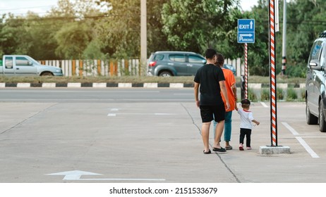 Back View Of Asian Family On The Way Back To Car Parking Lot. Parents Take Care And Hold Their Young Son By Hand As They Walk Back To The Car At Outdoor Parking Lot. Family Care And Concern Concept.