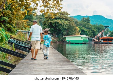 Back View Of Asian Grandfather And Grandchild Wear Protective Face Mask To Prevent Coronavirus (COVID-19) Walking In A Nature Path On Wooden Bridge Along River. Together Outdoors Family Concept.