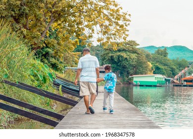 Back View Of Asian Grandfather And Grandchild Wear Protective Face Mask To Prevent Coronavirus (COVID-19) Walking In A Nature Path On Wooden Bridge Along River. Together Outdoors Family Concept.