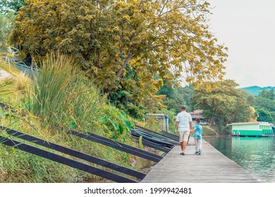 Back View Of Asian Grandfather And Grandchild Wear Protective Face Mask To Prevent Coronavirus (COVID-19) Walking In A Nature Path On Wooden Bridge Along River. Together Outdoors Family Concept.