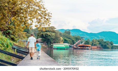 Back View Of Asian Grandfather And Grandchild Wear Protective Face Mask To Prevent Coronavirus (COVID-19) Walking In A Nature Path On Wooden Bridge Along River. Together Outdoors Family Concept.