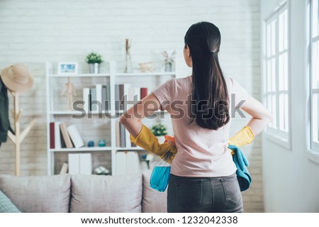 back view of asian housekeeper looking at the clean living room after she tidied up. young wife finished house chores putting hands in waist watching the bookshelf beside the sunlight window.