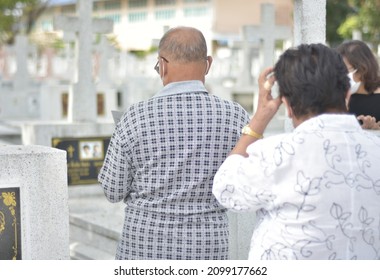 Back View Of Asian Senior Couple Standing For Mourn In Front Of The Grave To Remember The Deceased Relatives. Old Couple Standing For Prayer For The Deceased At Christ Cemetery. Selective Focus