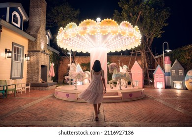 Back View Of Asian Woman In Dress Walking To Illuminated Carousel At Amusement Park In The Night