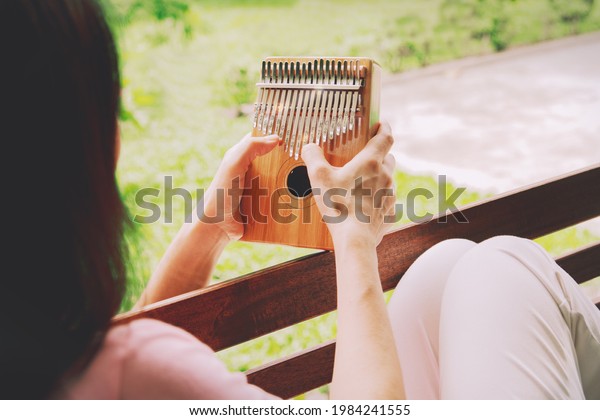 Back view Asian woman\
enjoys a kalimba, or thumb piano, an African traditional instrument\
in her garden : Woman enjoyed the melodious sound of the thumb\
piano playing.