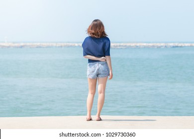 Back view of Asian woman traveler in blue casual and jean shorts standing on the stone terrace and looking around the sea ocean.