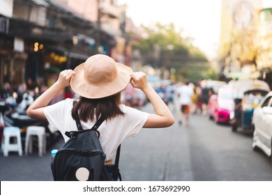 Back view of asian young woman traveller at street market. Hand hold a hat and smartphone. Backpack Traveling lifestyle in urban city on vacation summer. Relax outdoor at Khaosan, Bangkok, Thailand.