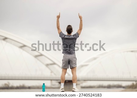 Back view of athletic young man in grey sportswear raising arms triumphantly outdoors. White modern bridge architecture creates minimalist backdrop while water bottle suggests post-workout celebration