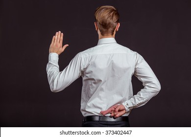 Back View Of Attractive Young Blond Businessman In White Classical Shirt Keeping One Hand Behind His Back With Crossed Fingers And Showing Pale Of Other Hand, Standing Against Dark Background