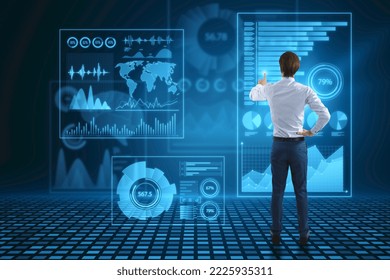 Back view of attractive young businessman using abstract glowing blue business charts and graphs hologram on dark background. Innovation, future and fintech concept