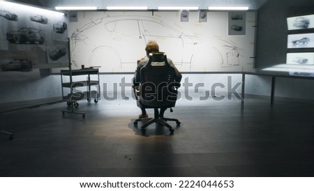Back view of automotive male designer looking at sketch blueprint of futuristic electric car future design on whiteboard. He sitting on chair in modern car design development studio.