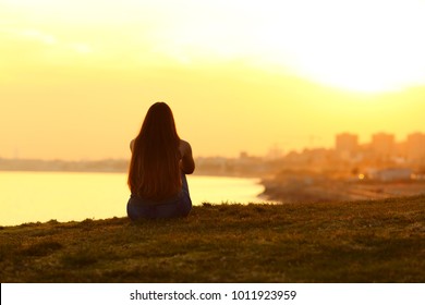 Back view backlight portrait of a single woman watching a sunset on the city with a warm light in the background