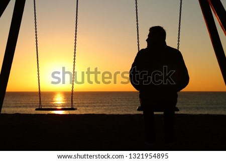 Back view backlighting silhouette of a man alone on a swing looking at empty seat at sunset on the beach in winter
