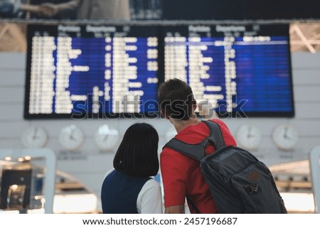 Back view. Backpacker couple checking boarding time at airport. Waiting in front of the arrival and departure board. Travel and transportation concept.