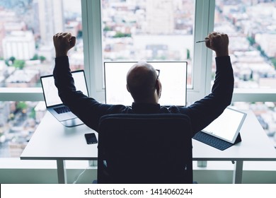 Back View Of Bald Excited Man Raising Fists Up Happy With Achievement Working With Gadgets At Desk Against Window In Office