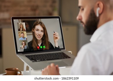 Back view of a bald male employee with beard who is listening to colleague on a video conference. A laptop screen view of the telecommunications application of an online meeting over a man's shoulder